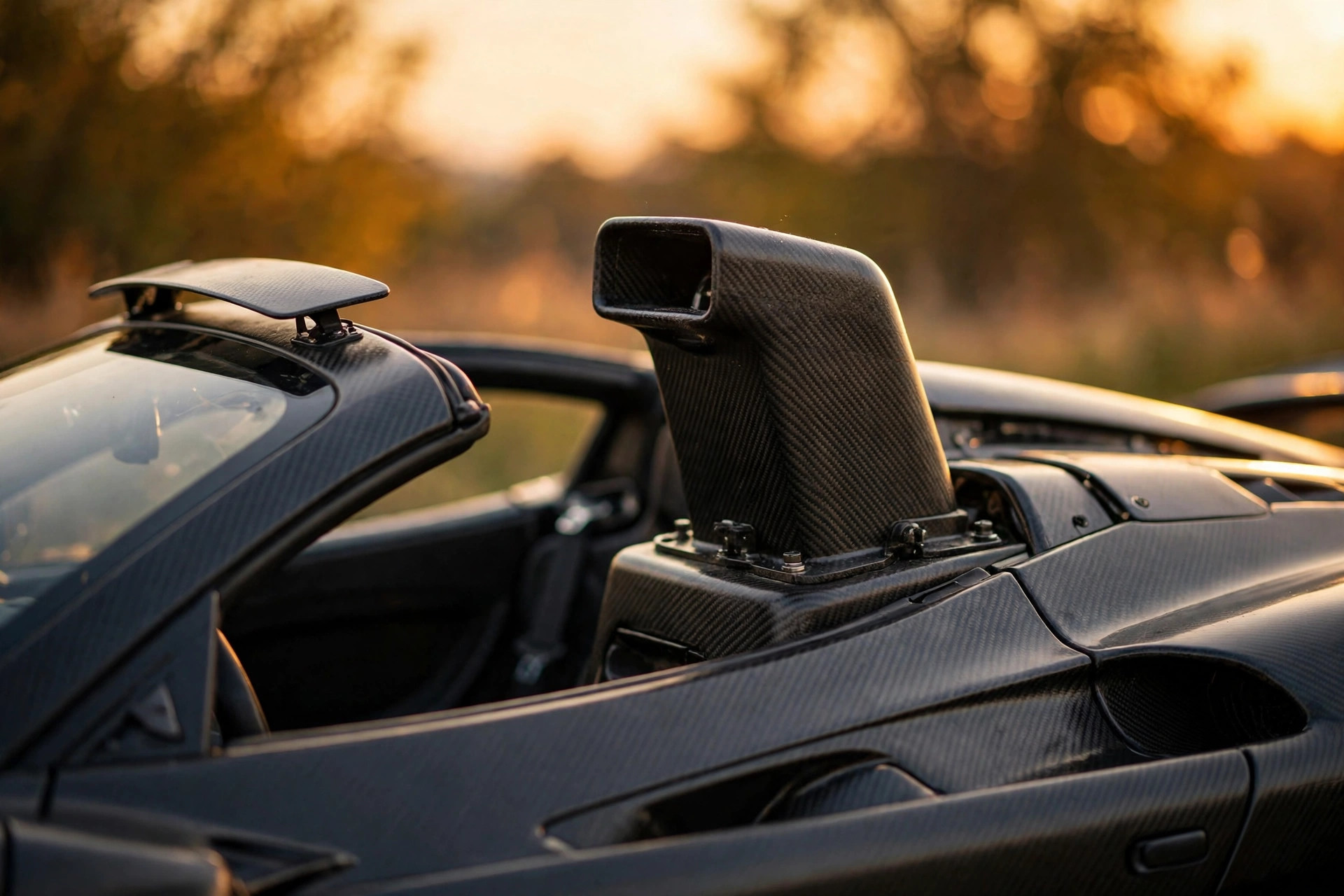 Editorial macro photograph in warm amber light of an open-top mid-engined supercar with its roof removed, showing the raised roof snorkel intake behind the cabin and a wind deflector at the top of the windscreen