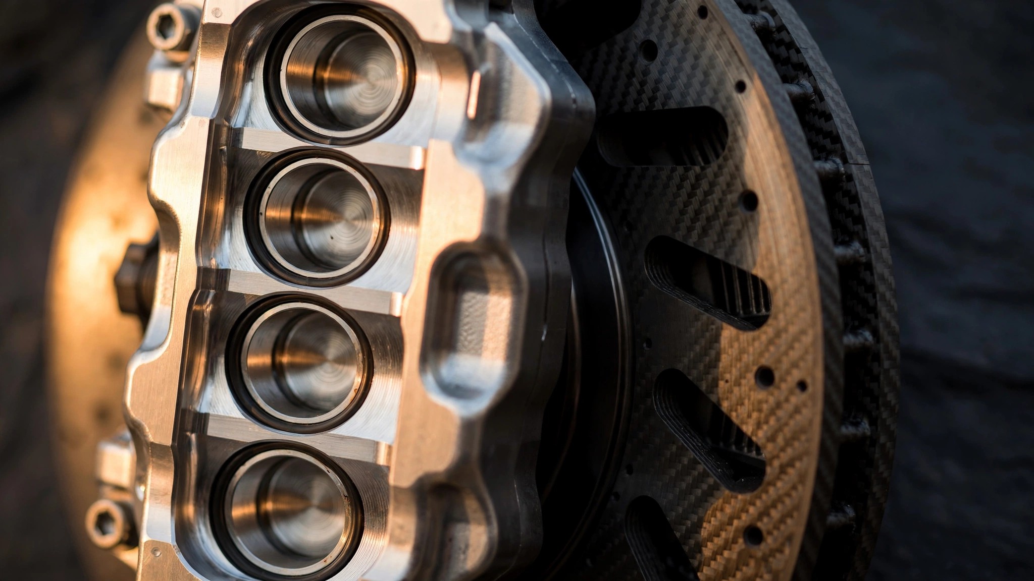 Macro photograph of a multi-piston monobloc brake caliper clamping a carbon ceramic rotor, warm directional light catching machined aluminum surfaces and ventilated rotor slots