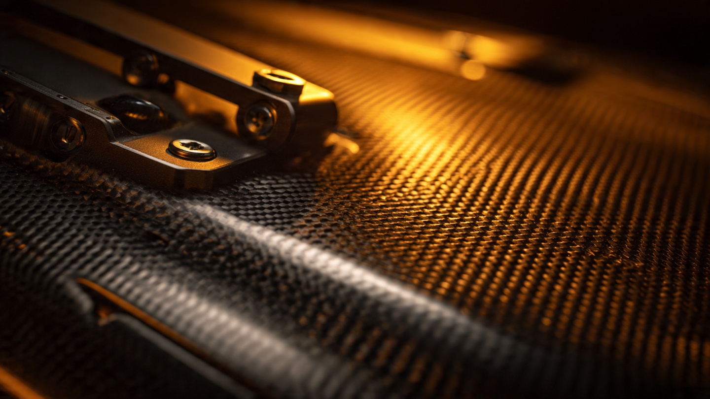 Macro photograph of carbon fiber weave and machined magnesium components from a convertible sports car roof mechanism under warm amber workshop lighting