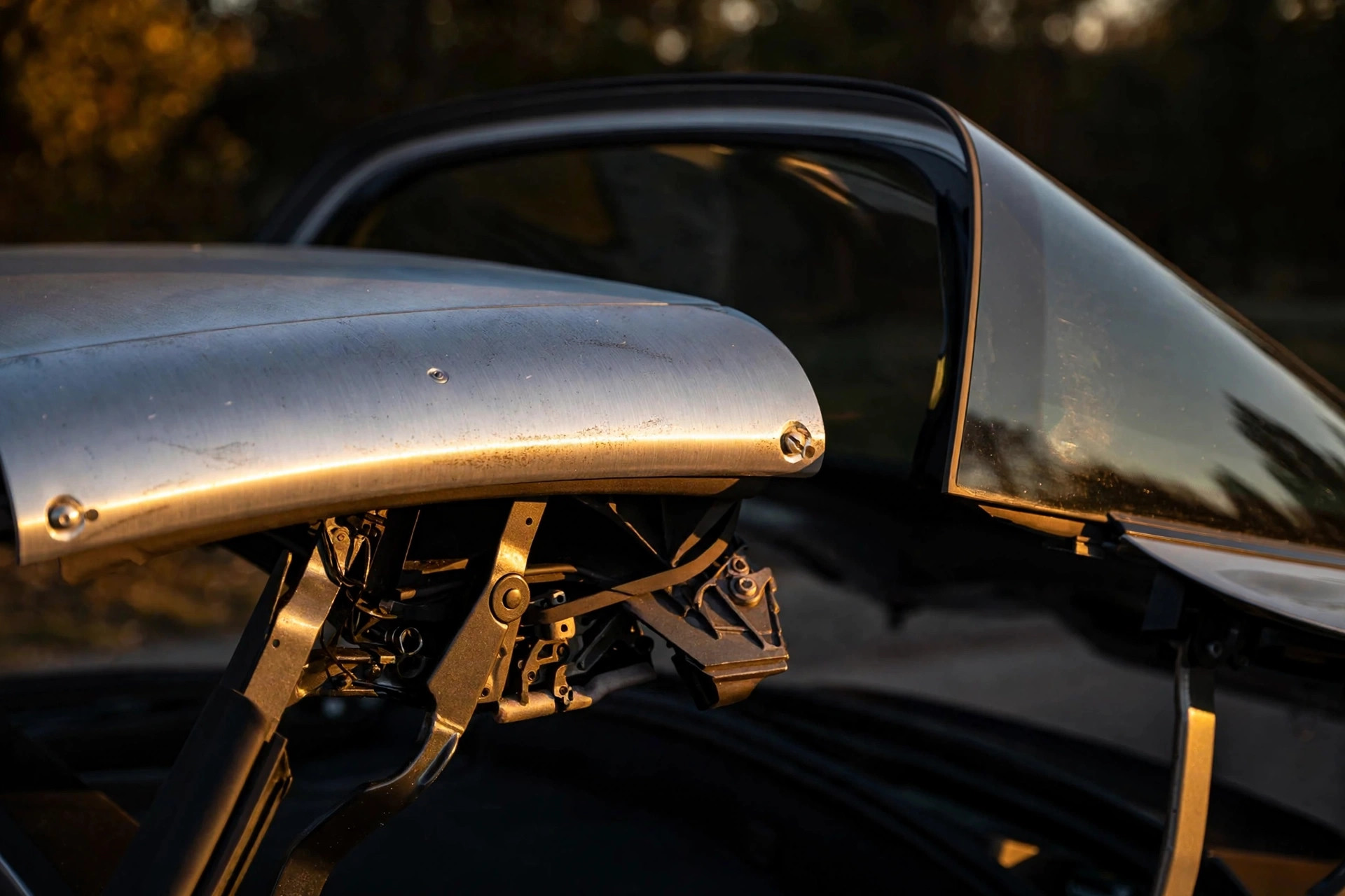 Editorial macro photograph in warm golden tones of a Porsche 911 Targa's silver roll bar and lifting rear glass at dusk, magnesium roof linkage visible mid-choreography, amber light on machined metal